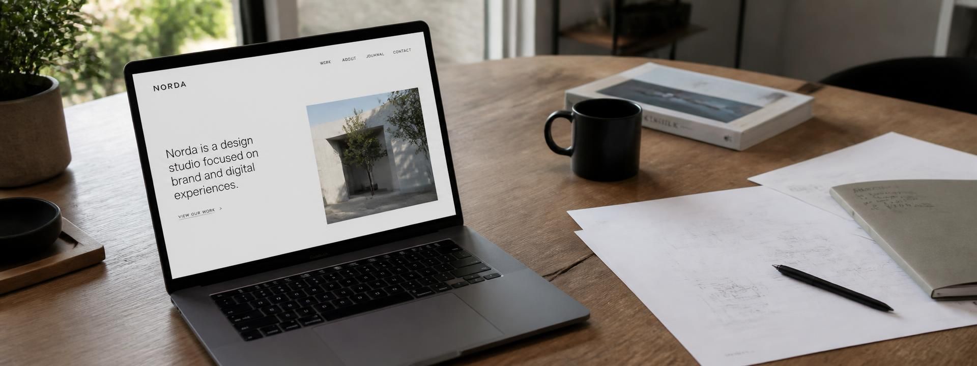 A laptop on a wooden desk showing a spacious minimal website, with papers and a mug beside it
